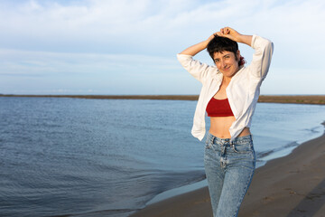 short-haired woman smiling on the beach, wearing white shirt, red top and denim, blue sky and calm sea.