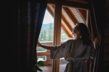 Young caucasian woman opening curtains in a wooden chalet cabin in the mountains. Comfortable rest in a log house in nature