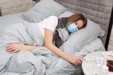 Sick woman in a medical mask takes a thermometer to check her temperature. Concept of fatigue from prolonged illness.