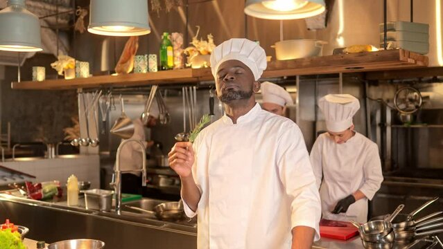 African-American male chef stands in restaurant professional kitchen and inhales good smell of fragrant herbal spices. Multicultural smiling cook selects ingredients for preparation of dish.