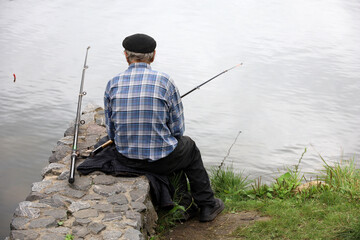 Fisherman sitting near the water with a fishing rod, rear view. Old man angling on the lake coast
