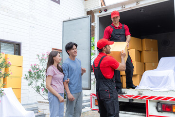 Asian couple newlywed move to new house using professional delivery logistic moving house business service, multiethnic Indian and Caucasian teamwork employees working uploading lifting cardboard box