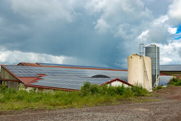 Farm buildings with rooves covered with solar panels and grain silos under a cloudy sky  view of solar panels on the rooves of barns and farm buildings. © Serhii