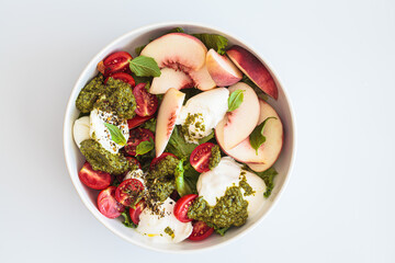 Summer burrata salad with peaches, cherry tomatoes, pesto and spices in bowl on white background.