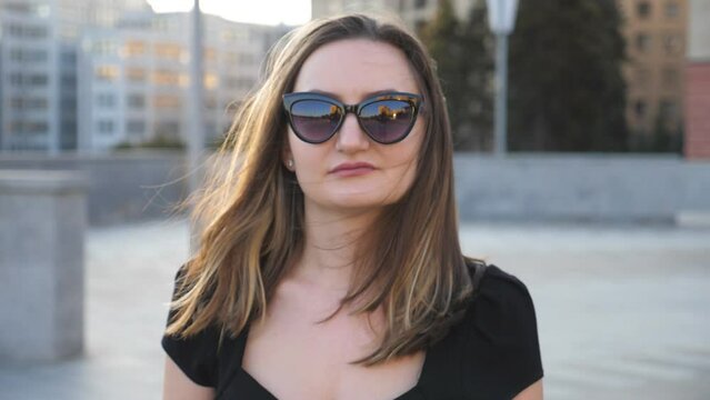 Portrait of young businesswoman in sunglasses walking in city street. Attractive business woman looking at camera. Face of confident girl commuting to work. Blurred background. Close up Slow motion