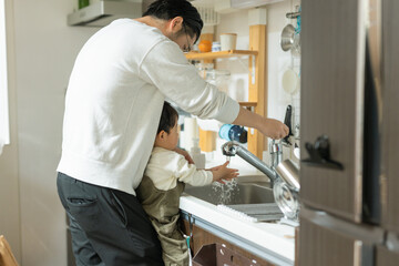 春の日中の台所で手を洗う日本人の小さな男の子と抱っこする父親 - obrazy, fototapety, plakaty