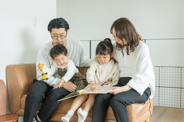 春の昼間、部屋のソファに座って本を指さす日本人の子どもと両親