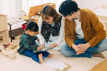 冬の日中、おもちゃが溢れるリビングで絵本を眺める日本人の子どもと父と母 - obrazy, fototapety, plakaty