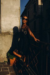 A young girl in the Italian patio at sunset in a low key. Little black dress with black glasses.