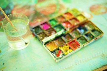 Old dirty watercolor paint set and glass of water with a brush on the table