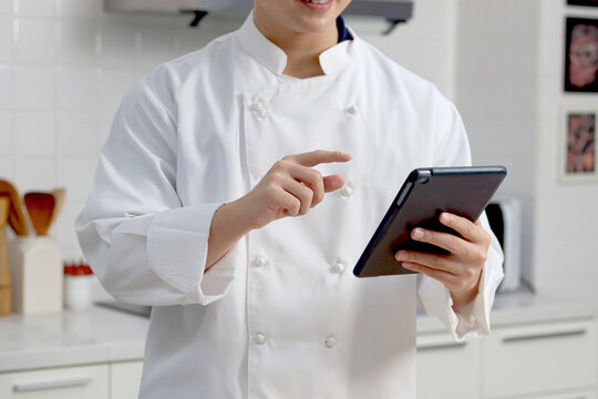Man in white chef uniform holding and pointing digital tablet, using tablet for searching tasty meal recipes online internet, man cooking meal food, young chef man cooking food at home.