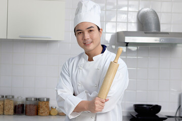 Happy smiling Asian man in white chef uniform with hat, holding wooden rolling kneading stick,...