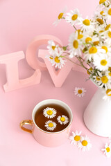 Bouquet of chamomiles, chamomile tea in a pink cup and three-dimensional pink letters LOVE on a pink background. Daisies in a cup. Photo