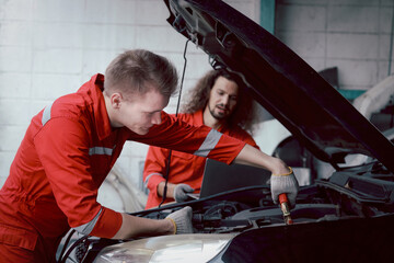 Mechanic man in red uniform charging car battery with electricity through jumper cables. coworker work together at vehicle garage, technician team repairing customer car automobile at service shop.
