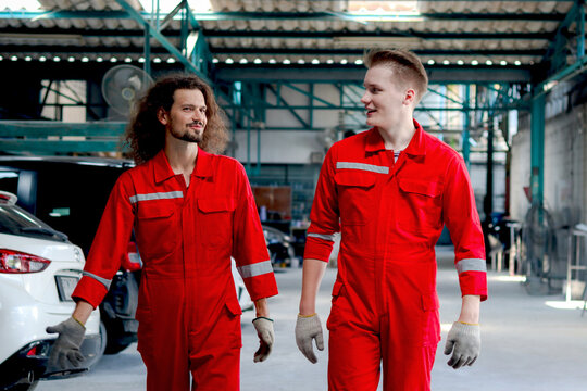 Two handsome mechanic men in red uniform walking through row of vehicle at garage, auto mechanic technician friend work together to repair and maintenance customer car automobile at car service shop.