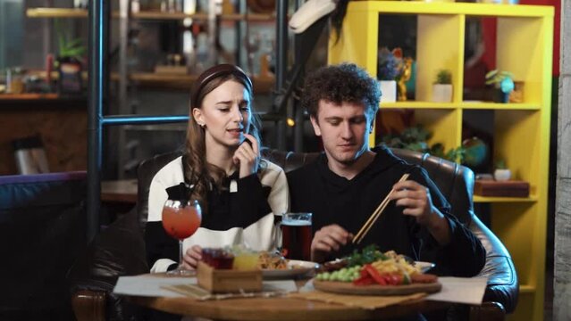 A girl and a boy are sitting in a restaurant they have ordered food and drinks. They don't like the food as they look very picky and displeased, with the boy reluctantly trying a bite.