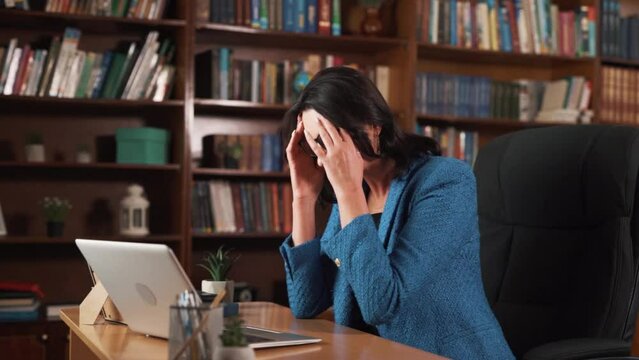 The woman is sitting at the table and working on her laptop. She looks tired, rubs her eyes, and touches her face with her hands. She leans back on the chair's backrest and relaxes.