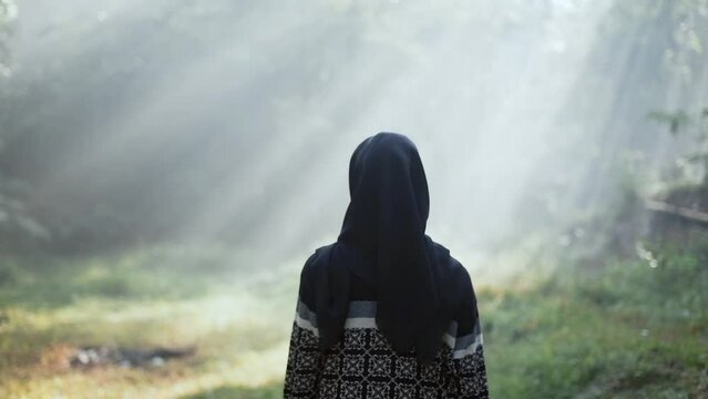 An Asian muslim hijab woman walking to the ray of light in the middle of a forest. View from the back