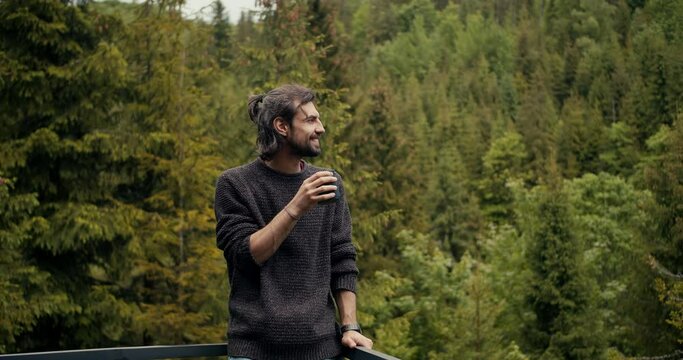 Overview of a happy brunette guy who is standing on the balcony, drinking tea and looking at the coniferous forest in ogres