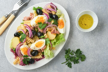 Tuna salad with pasta, eggs, potatoes, olives, red onions and sauce in white plate on old light gray concrete table background. Nicoise salad. French cuisine. Top view. Flat lay.