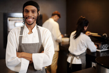 Portrait of cheerful restaurant sous-chef in white shirt and grey apron standing in kitchen