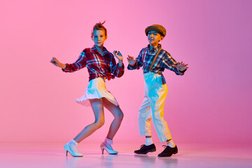 Fototapeta na wymiar Beautiful children, boy and girl in stylish retro clothes dancing against pink studio background in neon light. Concept of childhood, hobby, active lifestyle, performance, art, fashion