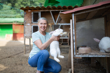 A farmer woman holds a white rabbit in her hands next to the cage.