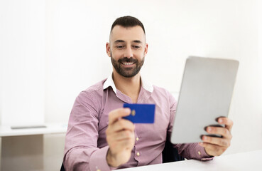 Happy caucasian mature man in shirt sits at table with tablet, uses credit card