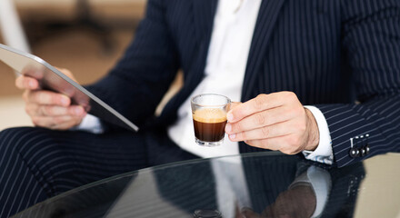 Confident caucasian mature man in suit drinking cup of coffee, uses tablet to manage business