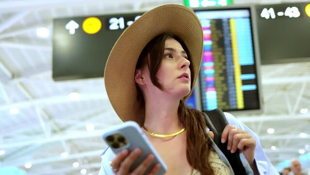 Charming young lovely Caucasian woman standing at the airport focused wearing hat and using smartphone online. Stunning charming girl texting in internet before flight. Travelling concept.