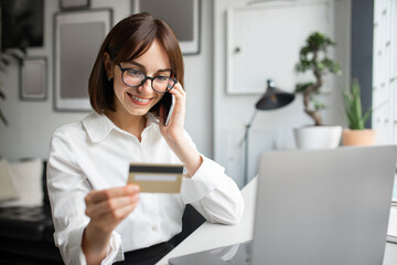 Businesswoman making mobile payments, holding credit card and talking on phone, sitting in cafe or coworking space