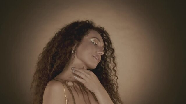 Attractive curly haired young woman with golden makeup smiling and looking to the side and touching her shoulder gently, golden paint and accessories, close up video.