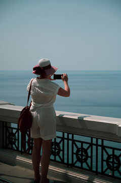 giovane donna con cappello e nastro ammira il mare dall'alto scattando foto dal suo smartphone in un contesto di serenit&agrave; e pace