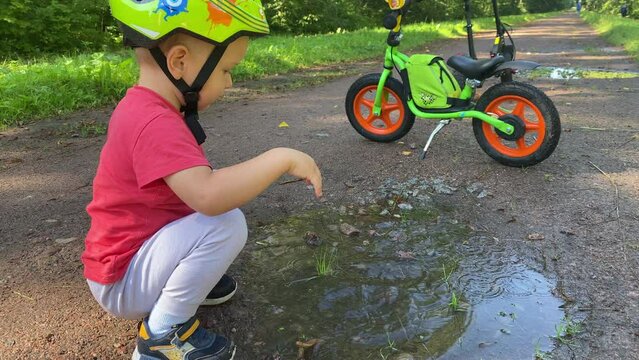 toddler child with balance bike in summer park, cute kid playing in puddle