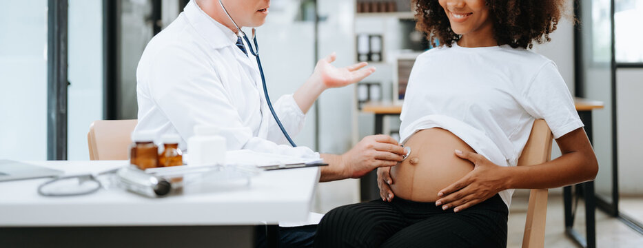 Pregnant african woman has appointment with doctor at clinic. Male gynaecologist OB GYN medic specialist with stethoscope listens to baby's heartbeat in mother's belly. Pregnancy, health care .