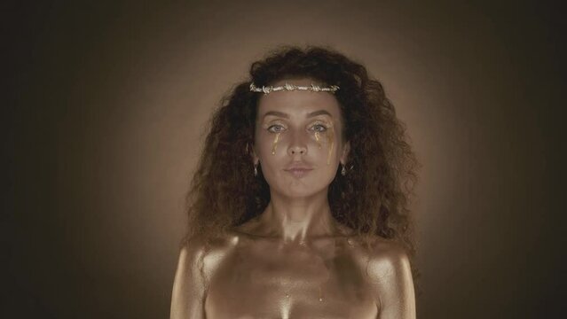 Attractive curly haired young woman with golden makeup smiling and looking at the camera, golden paint and accessories, crown of thornes on the head, close up video.