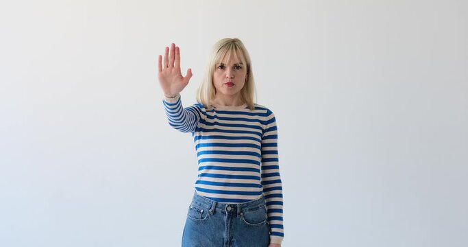 Unsmiling blonde woman is seen on a clean white backdrop, making the 'stop' gesture with her hand, showing her palm to signal halting or prohibiting something.