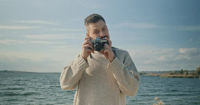Portrait of attractive happy bearded young man taking pictures with camera looking at camera smiling. Beautiful handsome male standing near lake enjoying views spending time outdoors.