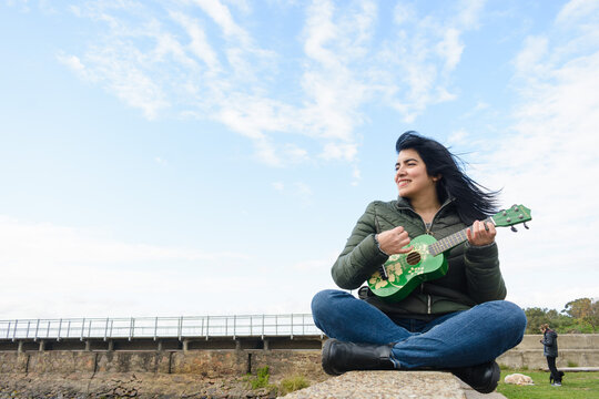 young latin woman sitting outdoors playing ukulele, out for walk enjoying weekend morning.