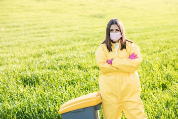 A woman in a yellow protective suit stands in the middle of a green field in a protective mask, wearing pink gloves. A woman stands near the garbage can and calls to save the planet
