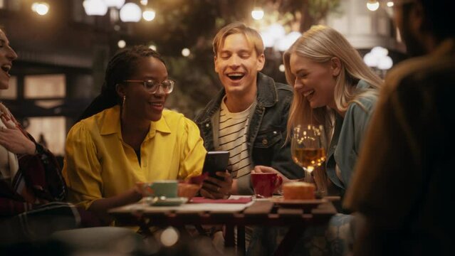 Young Female Sitting on a Terrace with Multiethnic Friends, Showing Holiday Photos on a Smartphone. Young Men and Women Gathered Together in a Cafe, Having Fun Thoughtful Conversations in the Evening