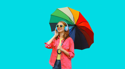 Happy cheerful laughing young woman with colorful umbrella listening to music in headphones wearing pink jacket on blue studio background