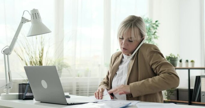 Female entrepreneur is depicted multitasking in her office. She is seen engaging in a phone conversation while briskly working with papers. With a sense of urgency, she efficiently manages both tasks.