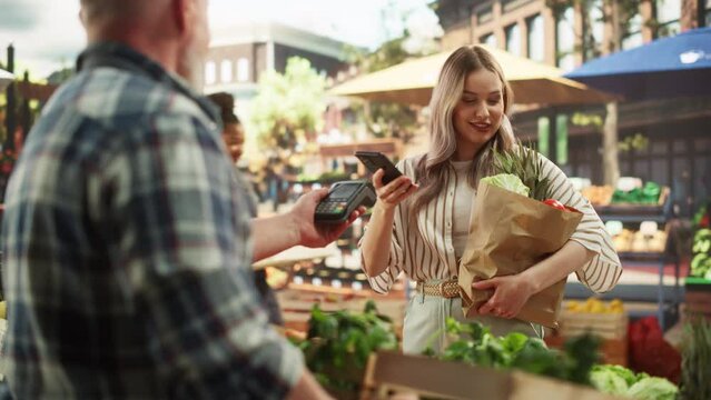 Modern Female Shopper Using Smartphone with Contactless Payment Technology to Pay for Organic Vegetables at a Farmers Market. Street Vendor Holding an Electronic Online Payment Terminal Device