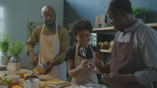 Senior African American grandfather giving whisks covered with whipped egg whites to grandson, kid licking them while baking together with family in kitchen at home