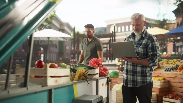 Successful Senior Farmer Working on a Laptop Computer, Writing Emails to Suppliers to Orders Produce for His Organic Street Vendor Stall with Natural Fruits and Vegetables