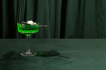 Green cocktail in a crystal glass with a sugar cube on a silver spoon on a dark green background