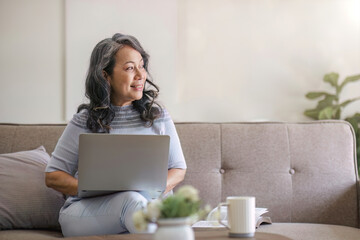 Happy Asian 60s retired woman enjoys social media on laptop while relaxing in living room.