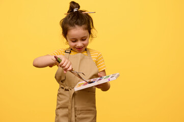 Smiling little child girl artist, schoolgirl dressed as an artist in beige apron, learning painting...