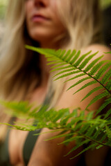 Naklejka premium beautiful blonde woman in green bikini blurred in front of green fern, focus on fern, outside shooting, natural